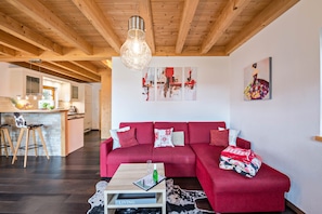 Un salon moderne avec un canapé rouge, des poutres en bois, un luminaire suspendu et une cuisine en arrière-plan.