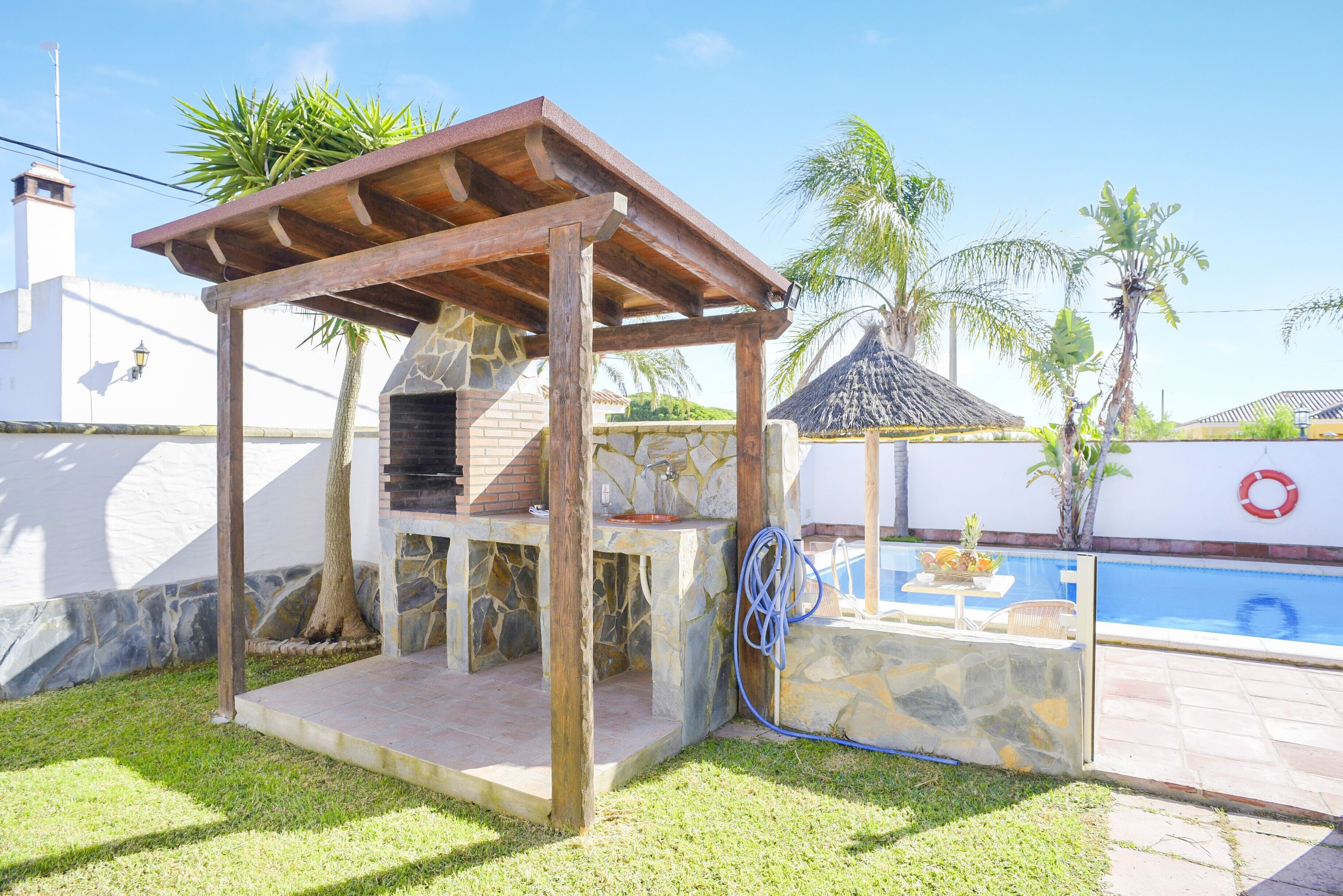 A barbecue area with stone walls, a wooden pergola, and a pool in the background.