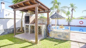 A barbecue area with stone walls, a wooden pergola, and a pool in the background.