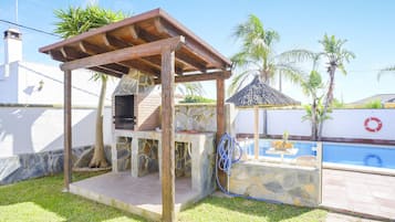 A barbecue area with stone walls, a wooden pergola, and a pool in the background.