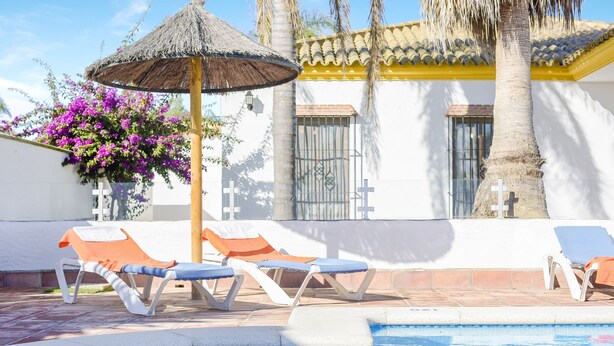A pool area with lounge chairs, a thatched umbrella, and a building with a tiled roof and palm trees.