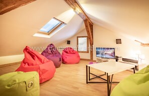 Quarto de sótão com teto inclinado, vigas de madeira, claraboia, televisão de tela plana, mesa e poltronas de espuma coloridas.