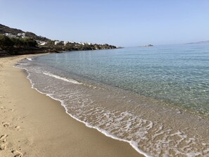 A beach with clear water, sandy shore, and buildings in the background.