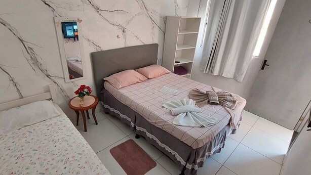 A bedroom with a bed, a bedside table with a vase of flowers, a mirror, and a wardrobe. - Guest House Palmeiras (Cabo Frio)