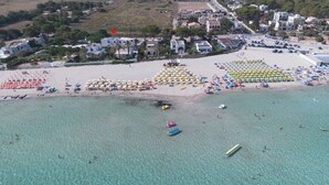 An aerial view of a beach with rows of umbrellas and beach chairs, a clear blue sea, and buildings in the background. - Villa Blumarine (San Vito Lo Capo)