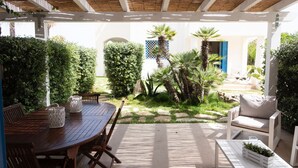 An outdoor patio with a wooden table and chairs, surrounded by lush greenery and palm trees. - Villa Blumarine (San Vito Lo Capo)