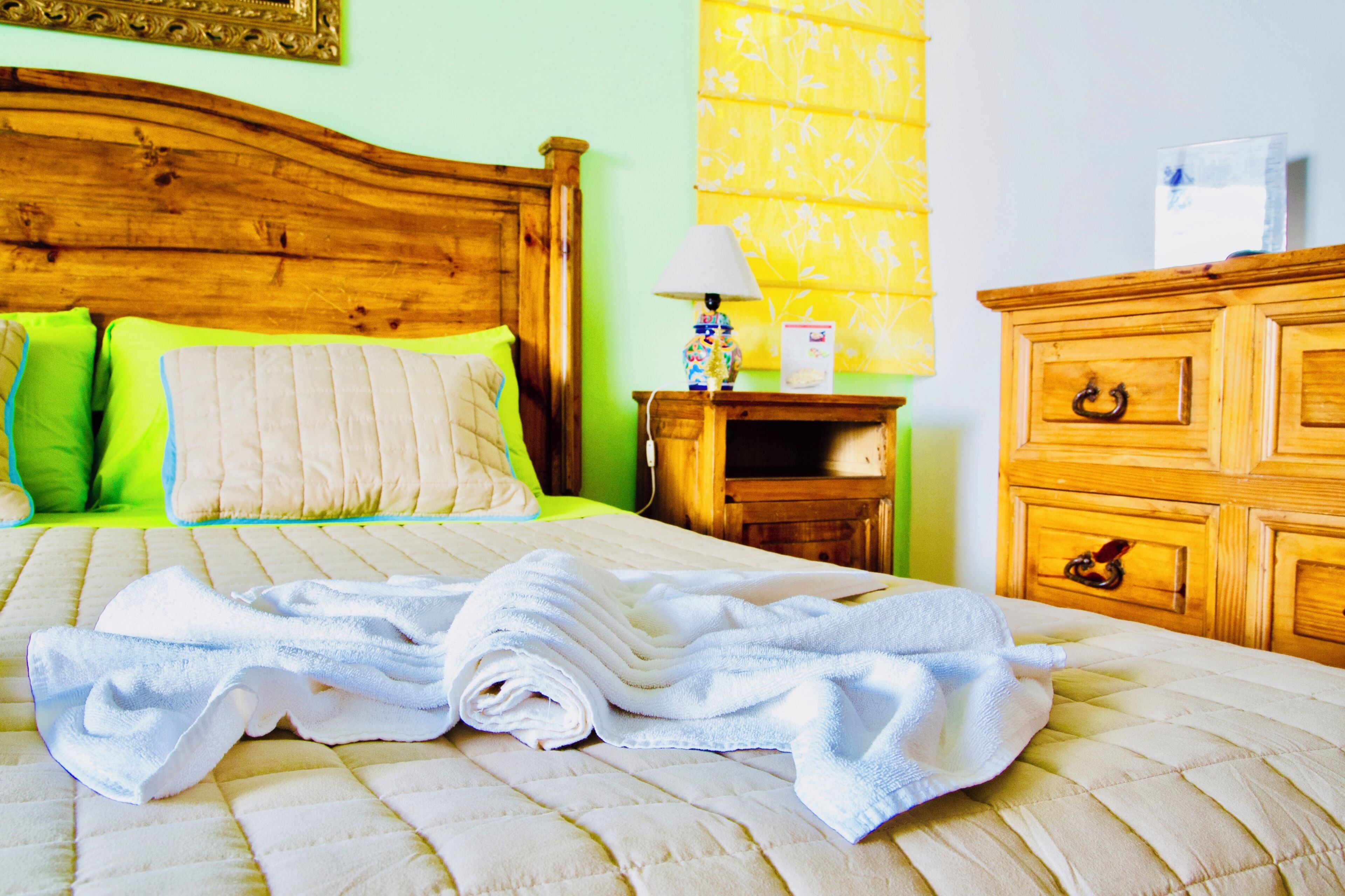 A neatly made bed with a rolled white towel, a wooden headboard, and a bedside table with a lamp and a box.