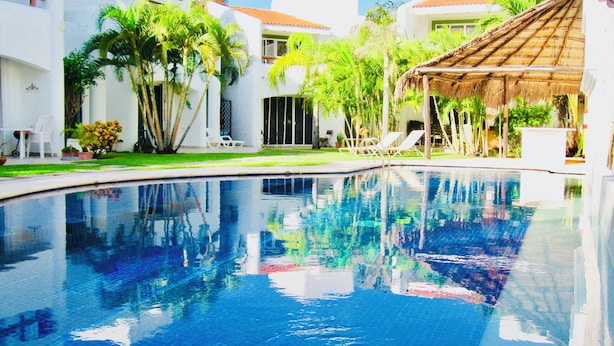A swimming pool with clear blue water, surrounded by white buildings with red-tiled roofs, and palm trees. - Room-playacar - Deluxe Family Comfort Ba (Playa del Carmen)