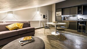 Un salón moderno con sofá, reposapiés y una mesa de comedor con sillas. Se ve una cocina con armarios oscuros y una encimera al fondo.