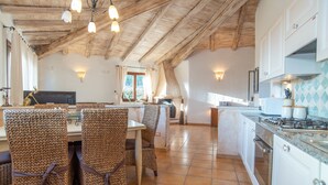 Une cuisine avec des poutres en bois, un coin repas avec des chaises en osier et un sol carrelé.