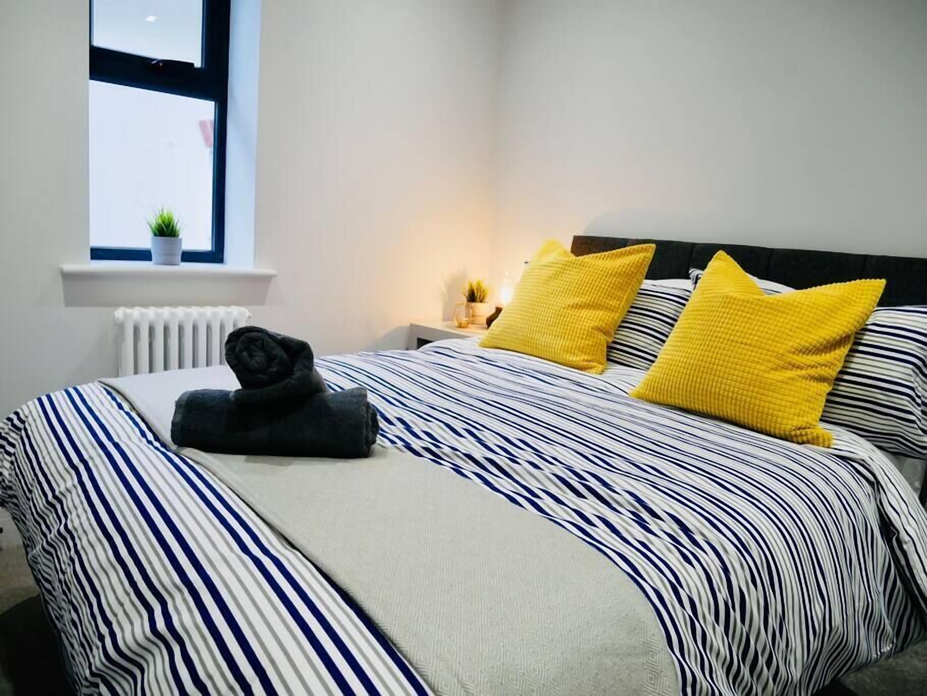 Ein ordentlich bezogenes Bett mit gestreifter Bettwäsche und zwei leuchtend gelben Kissen, ein kleiner Blumentopf auf einem Fensterbrett und ein Nachttisch mit einer Lampe.