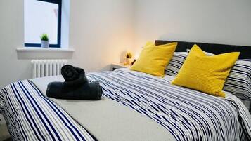 Ein ordentlich bezogenes Bett mit gestreifter Bettwäsche und zwei leuchtend gelben Kissen, ein kleiner Blumentopf auf einem Fensterbrett und ein Nachttisch mit einer Lampe.