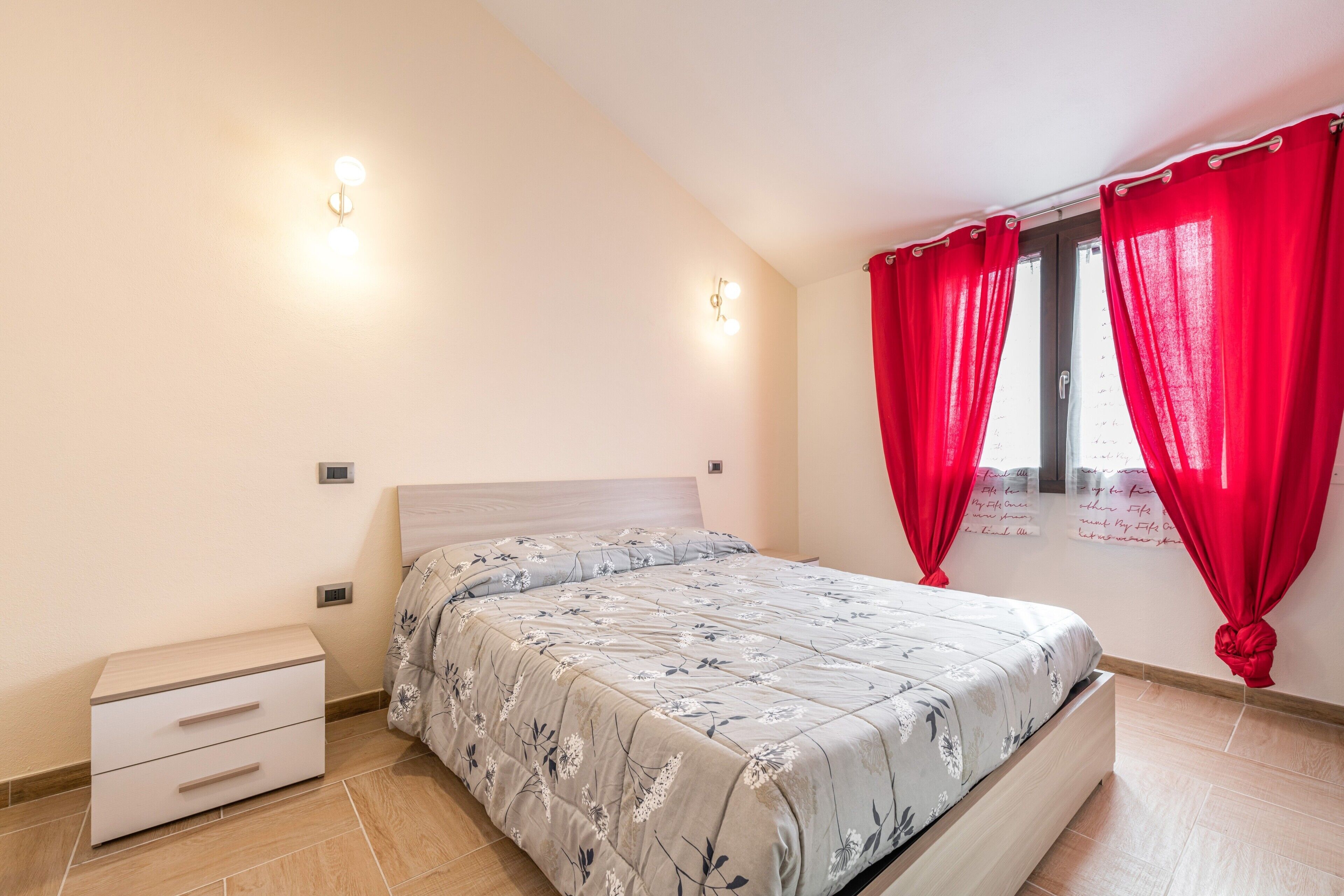 Een slaapkamer met een bed, een nachtkastje, een raam met rode gordijnen en wandlampen.