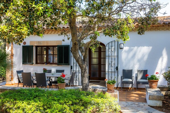 A white building with a tiled roof, a large tree, and outdoor seating with chairs and tables.