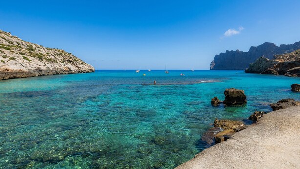 Un paysage côtier avec une eau turquoise claire, un terrain rocheux et des bateaux visibles au loin.