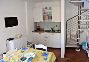 Une cuisine compacte avec des meubles blancs, un téléviseur encastré, une table à manger recouverte d’une nappe jaune et un escalier en colimaçon.