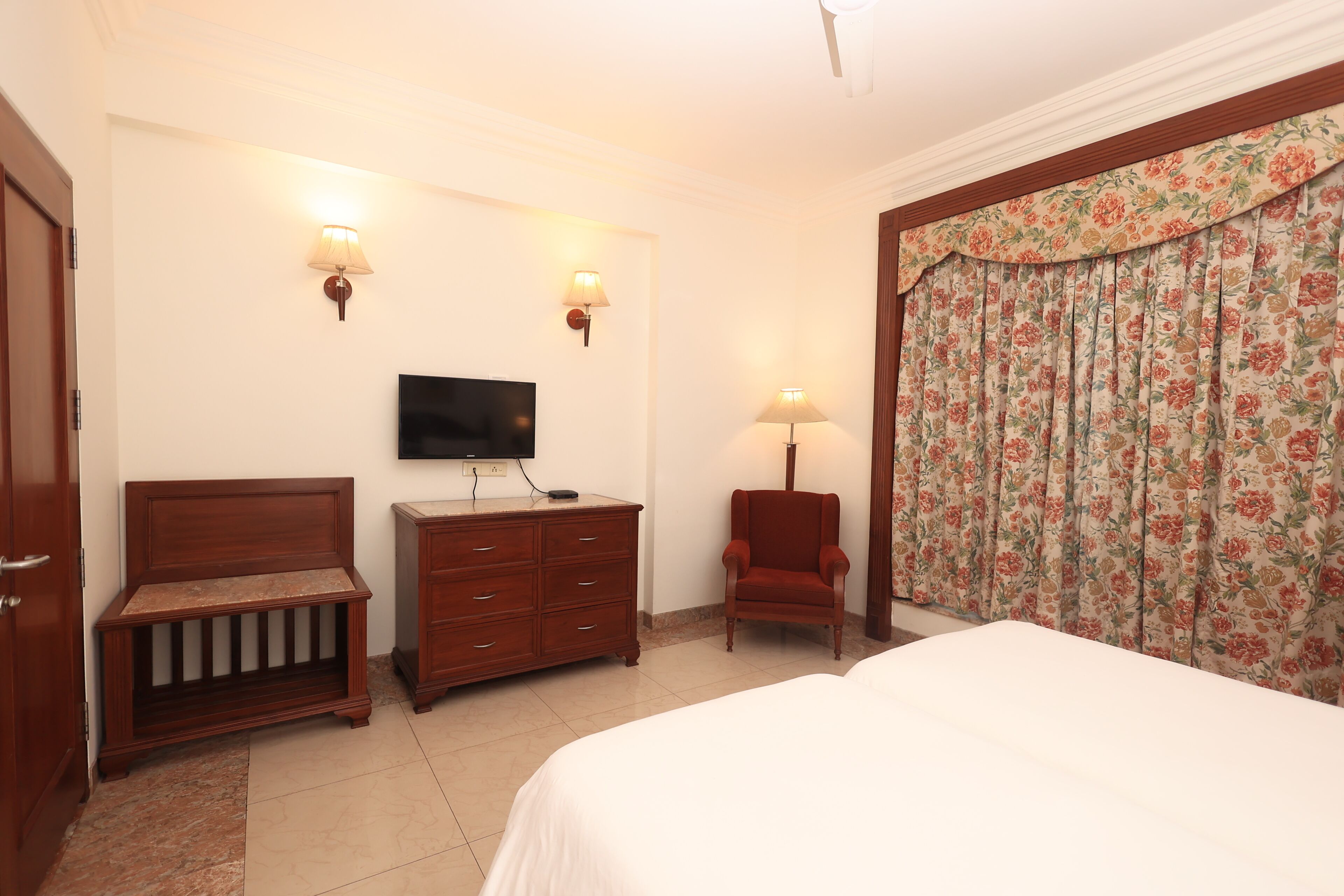A hotel room with a bed, a wooden bench, a dresser, a chair, a television, and a floral curtain.