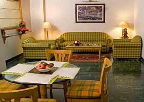 A cozy living room with a green plaid sofa set, a coffee table, and a dining table with chairs. There is a painting on the wall and a lamp on each side of the sofa.