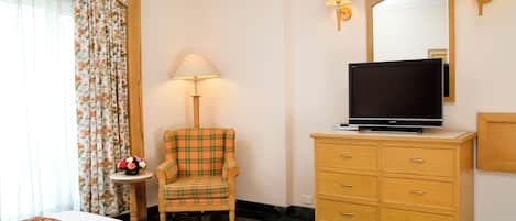 Habitación de hotel con cama, televisión sobre una cómoda de madera, un sillón a cuadros, una mesita con lámpara y cortina estampada.