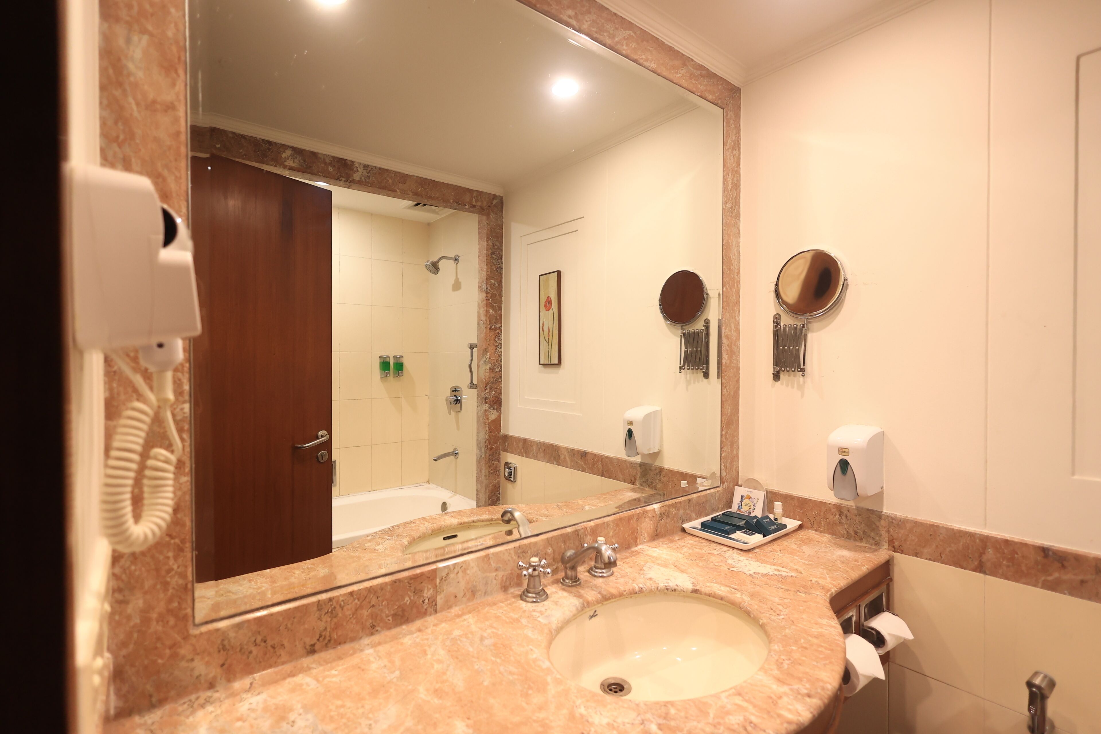 A bathroom with a marble sink, a large mirror, a handheld mirror, a towel dispenser, and a hand dryer.