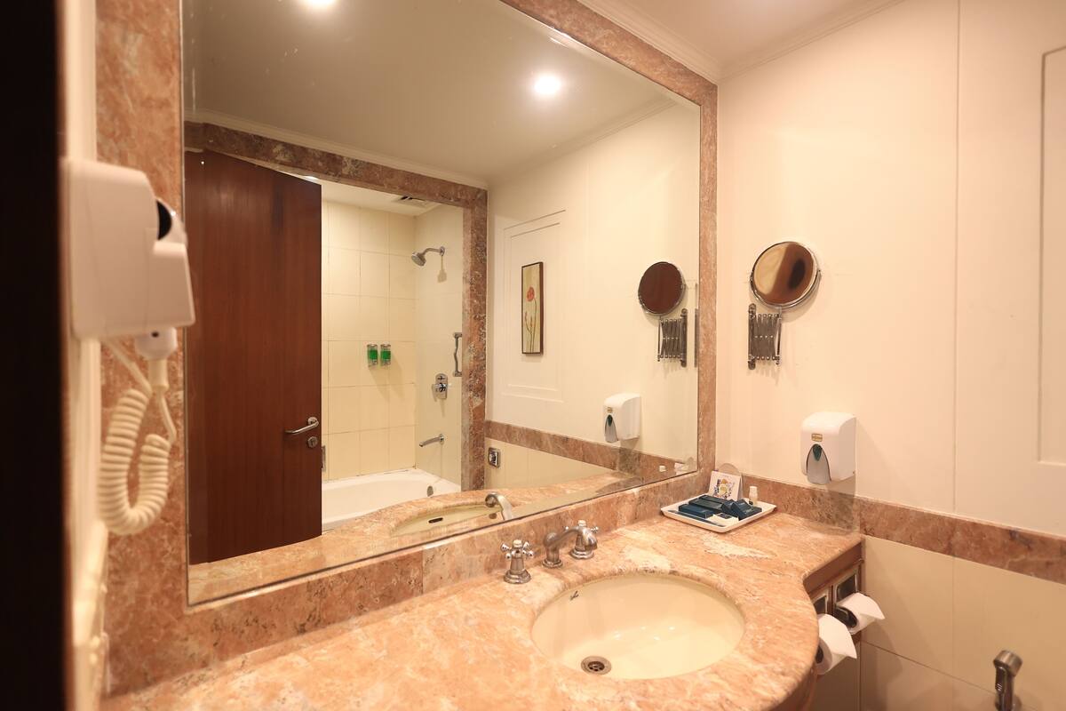A bathroom with a marble sink, a large mirror, a handheld mirror, a towel dispenser, and a hand dryer.