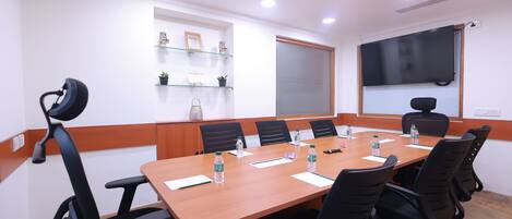 Sala de conferências com uma mesa comprida, cadeiras pretas, garrafas de água e uma tela de projetor.