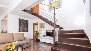 A modern living room with a brown staircase, a sofa, a coffee table with a vase of flowers, and a flat-screen TV on a white cabinet.