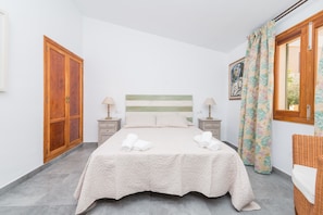 A bedroom with a bed, two bedside tables, a wooden wardrobe, a window with curtains, and a wicker chair.