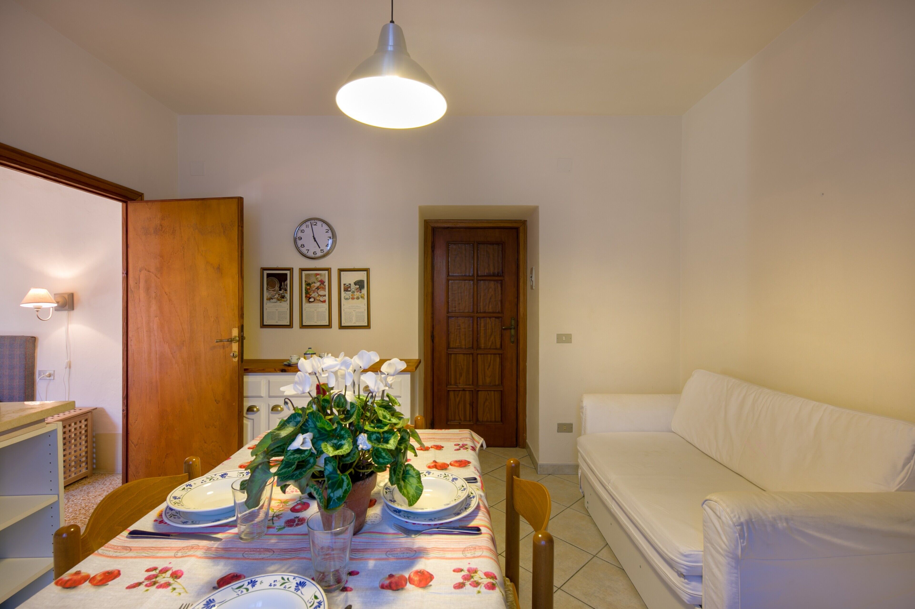A dining area with a table set for two, a wooden door, a white sofa, and a wall clock.