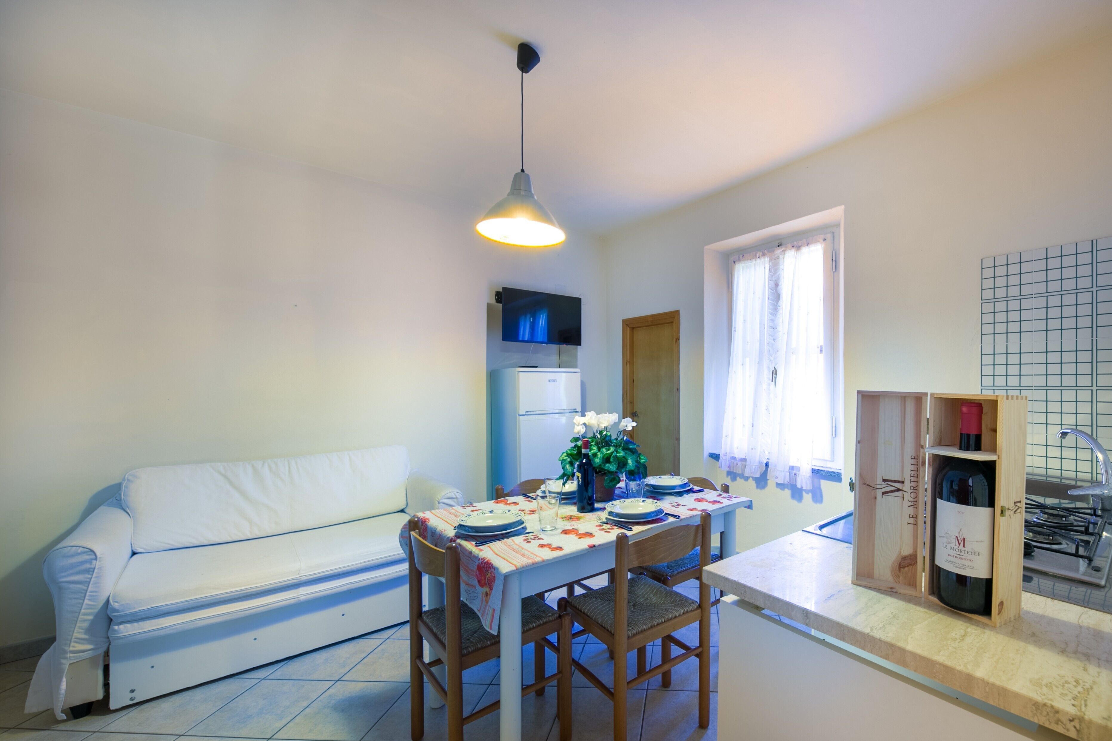 Ein Essbereich mit Tisch für vier Personen, einem weißen Sofa, einer Hängelampe, einem Fernseher über dem Tisch und einer Weinflasche in einer Holzkiste.