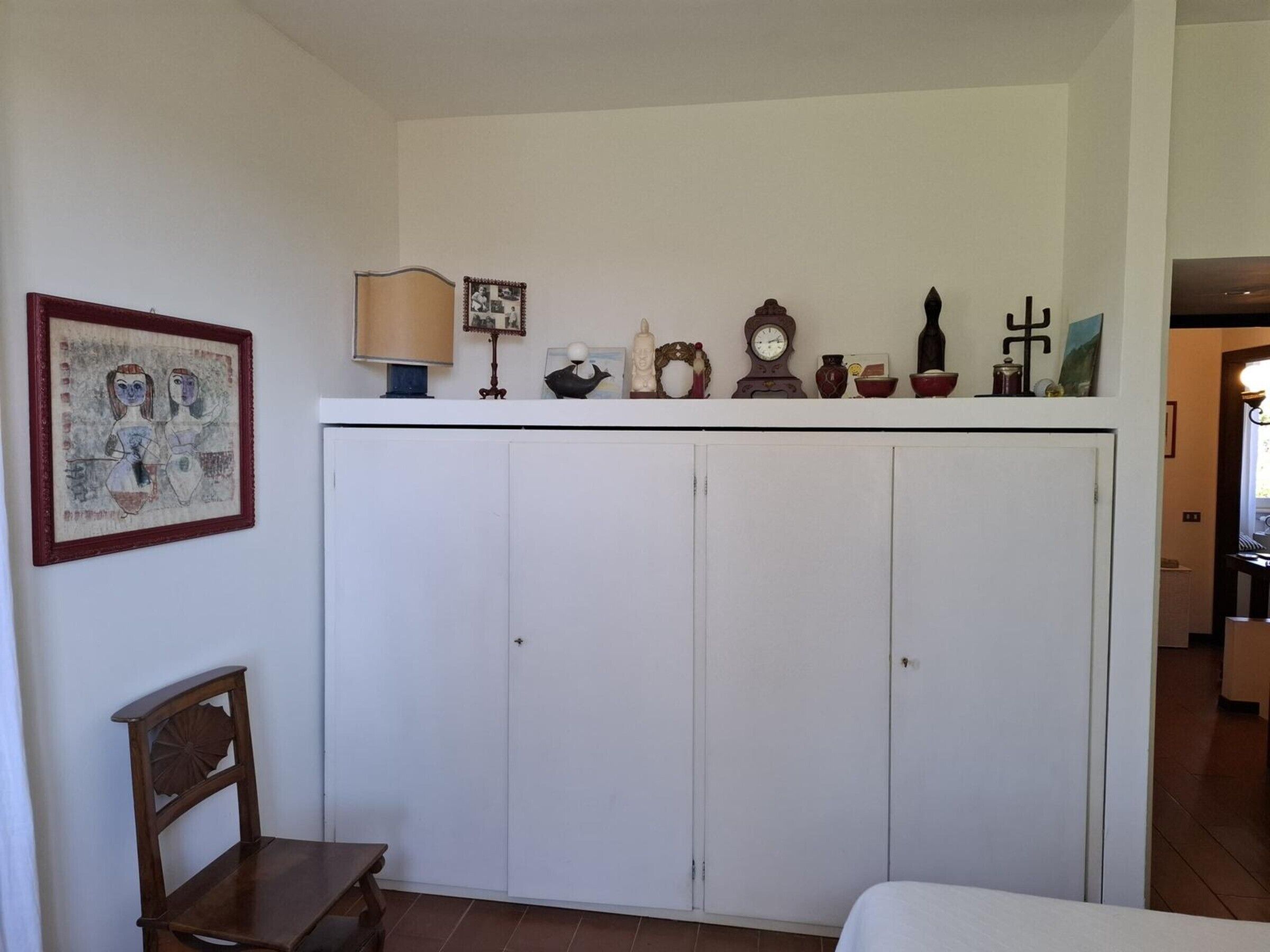 Una habitación con un armario blanco, una silla de madera, una cama y varios objetos decorativos en la pared.