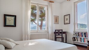 A bedroom with a bed, a window with curtains, a radiator, a wooden chair, and a bookshelf.