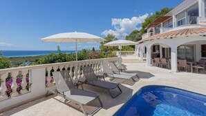 A poolside area with lounge chairs and umbrellas, overlooking a scenic view of the sea and greenery. - Stunning villa with pool & sea view – Villa Maitreya (san jaime, son bou, Alaior)