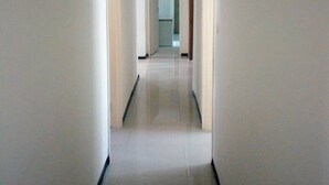 Un pasillo estrecho y bien iluminado, con paredes blancas y suelo de baldosas, que conduce a una habitación con una puerta al final.