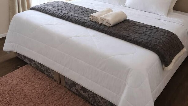 Una cama bien hecha con ropa blanca y una manta gris, un tapete rosa y una ventana con cortinas color beige.