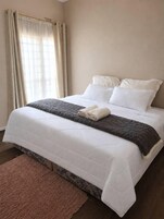 A neatly made bed with white bedding and a gray blanket, a pink rug, and a window with beige curtains.