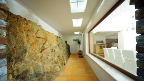 Un estrecho pasillo con paredes de piedra, suelo de baldosas y una ventana con sillas blancas.