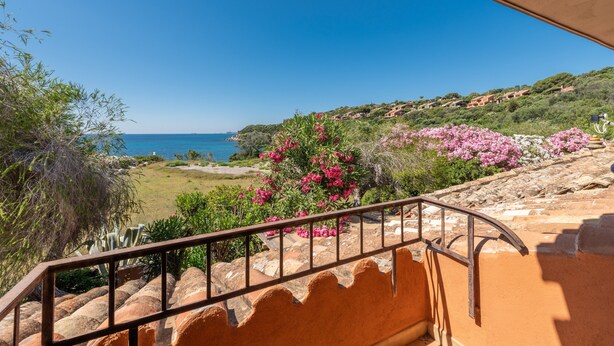 Uma varanda com vista para o mar, um jardim com flores cor-de-rosa e uma colina com edifícios.