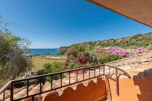 A balcony with a view of the sea, a garden with pink flowers, and a hillside with buildings.