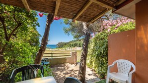 Uma área de estar coberta ao ar livre com vista para o mar, uma mesa, cadeiras e um muro coberto por plantas trepadeiras.
