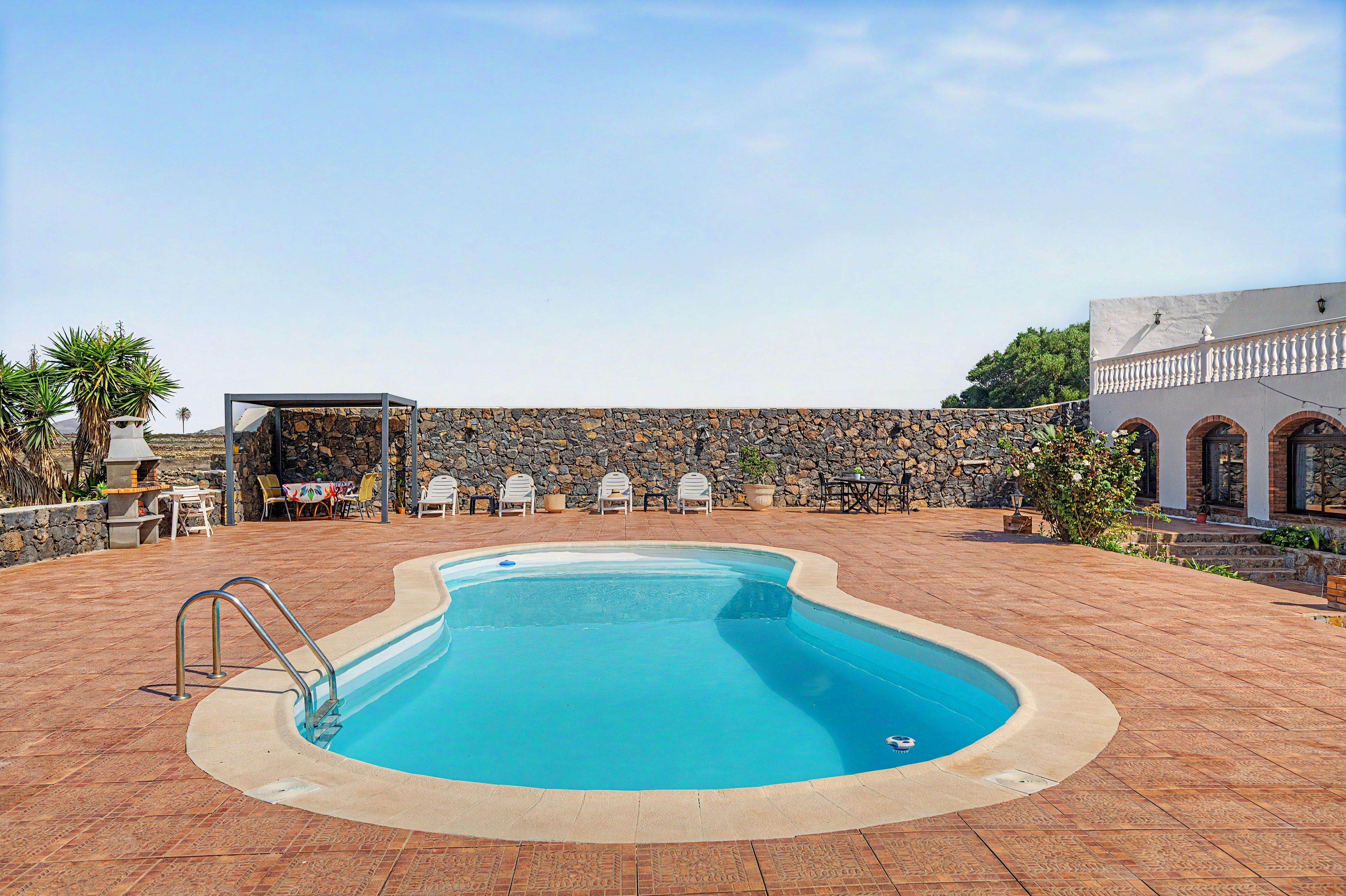 A swimming pool with a stone wall and chairs, surrounded by a patio area.