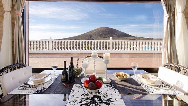 Une table dressée avec des assiettes, des couverts, un saladier de fruits, des bouteilles de vin et des verres, avec une vue sur une montagne par la fenêtre.