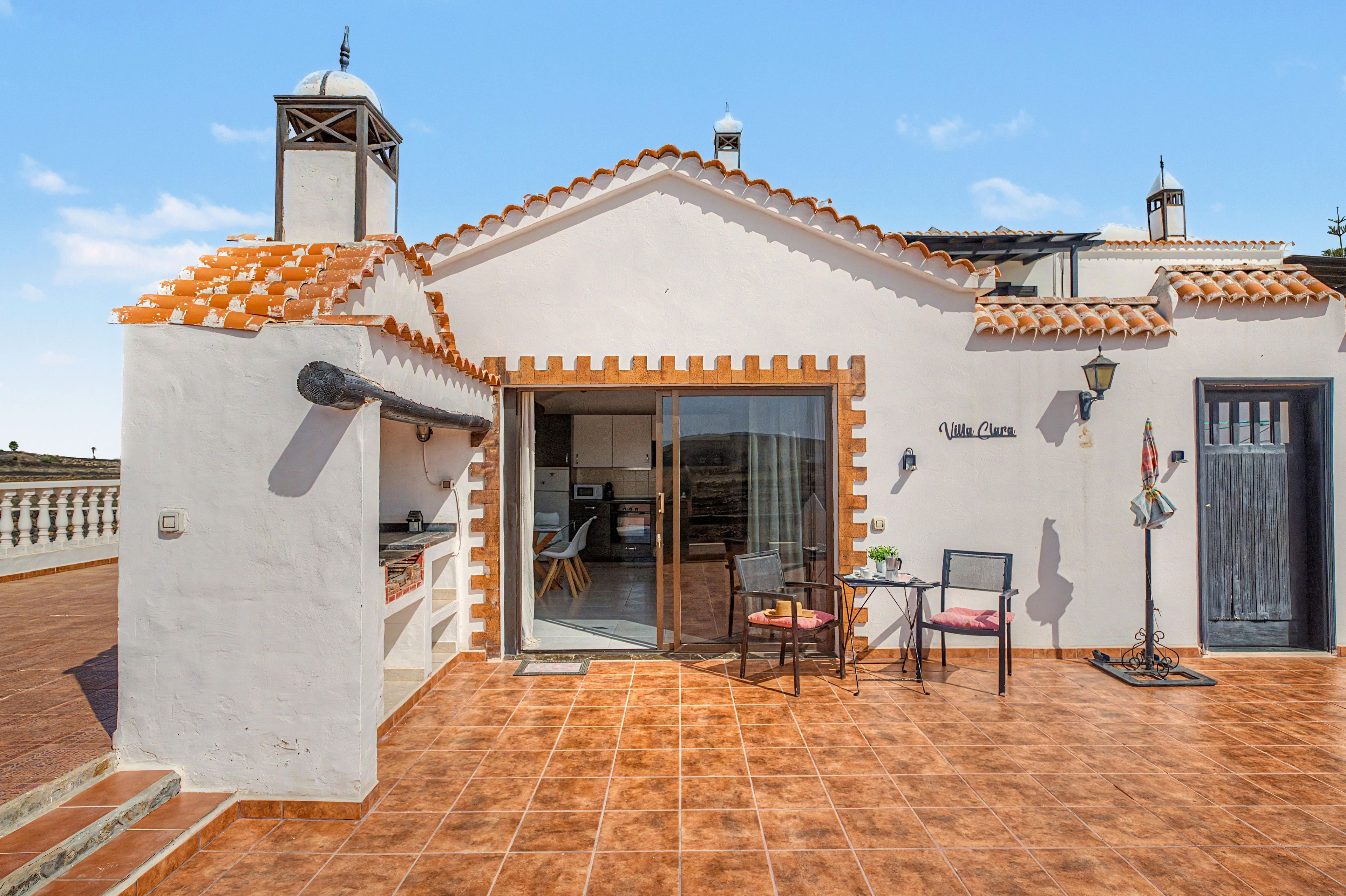A villa with a tiled roof, a dining area with a table and chairs, and a kitchen visible through a glass door.