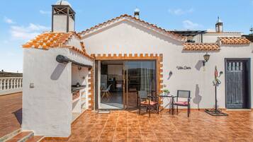 A villa with a tiled roof, a dining area with a table and chairs, and a kitchen visible through a glass door.