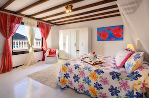 A bedroom with a bed, a wardrobe, a chair, a window with curtains, and a painting on the wall. - Clara Suite 3 with Large Pool, Stunning Views, Terrace and Garden (Lanzarote)