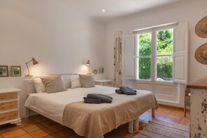 Un dormitorio con cama, mesitas de noche, lámpara, ventana con cortinas y vistas al exterior.