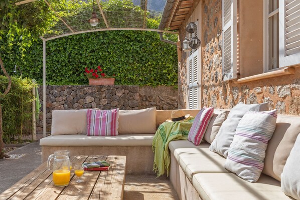Un espace extérieur accueillant, avec un mur en pierre, un banc rembourré et une table en bois sur laquelle sont posés une carafe de jus d’orange et des livres.