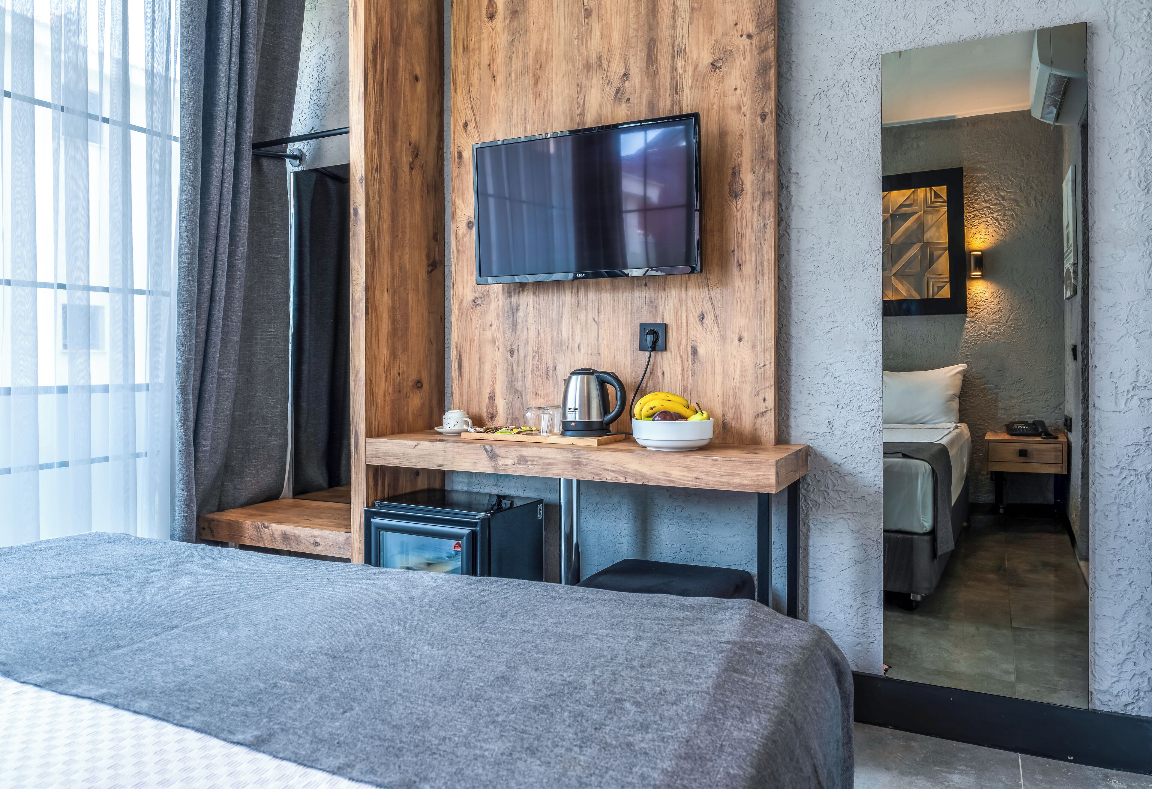 Quarto de hotel moderno com cama, TV fixada numa parede de madeira, uma pequena cozinha e banheiro visível por uma porta aberta.