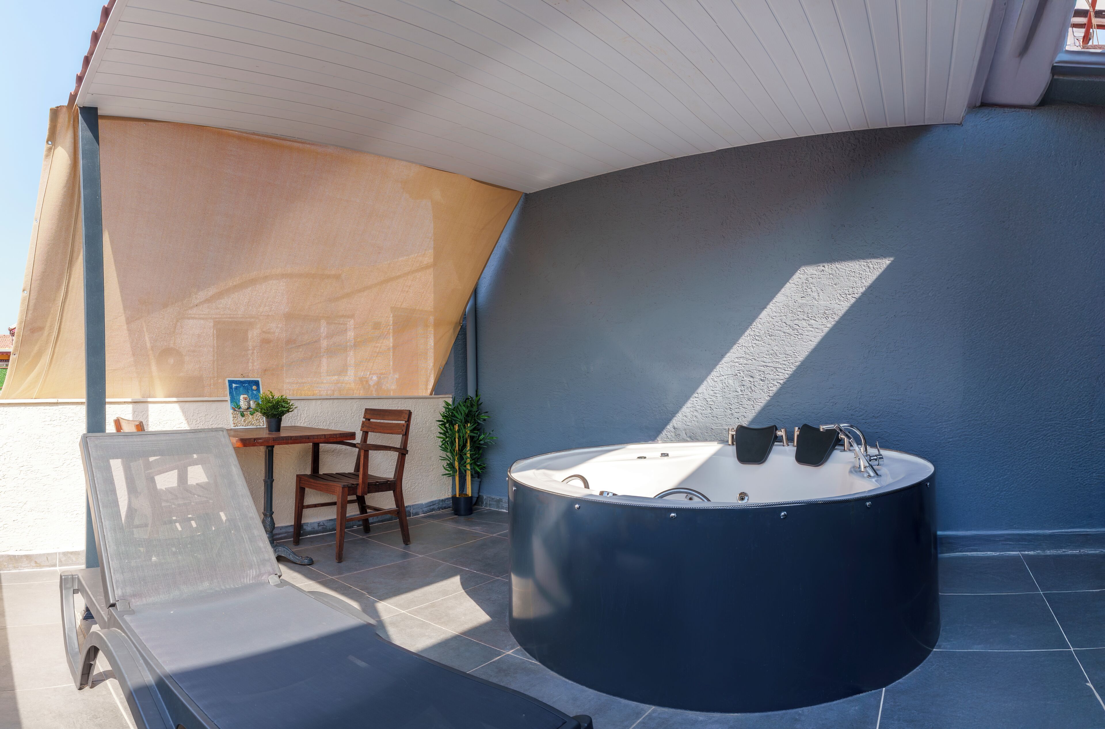 A sunlit outdoor area with a hot tub, a lounge chair, a table with chairs, and a potted plant.