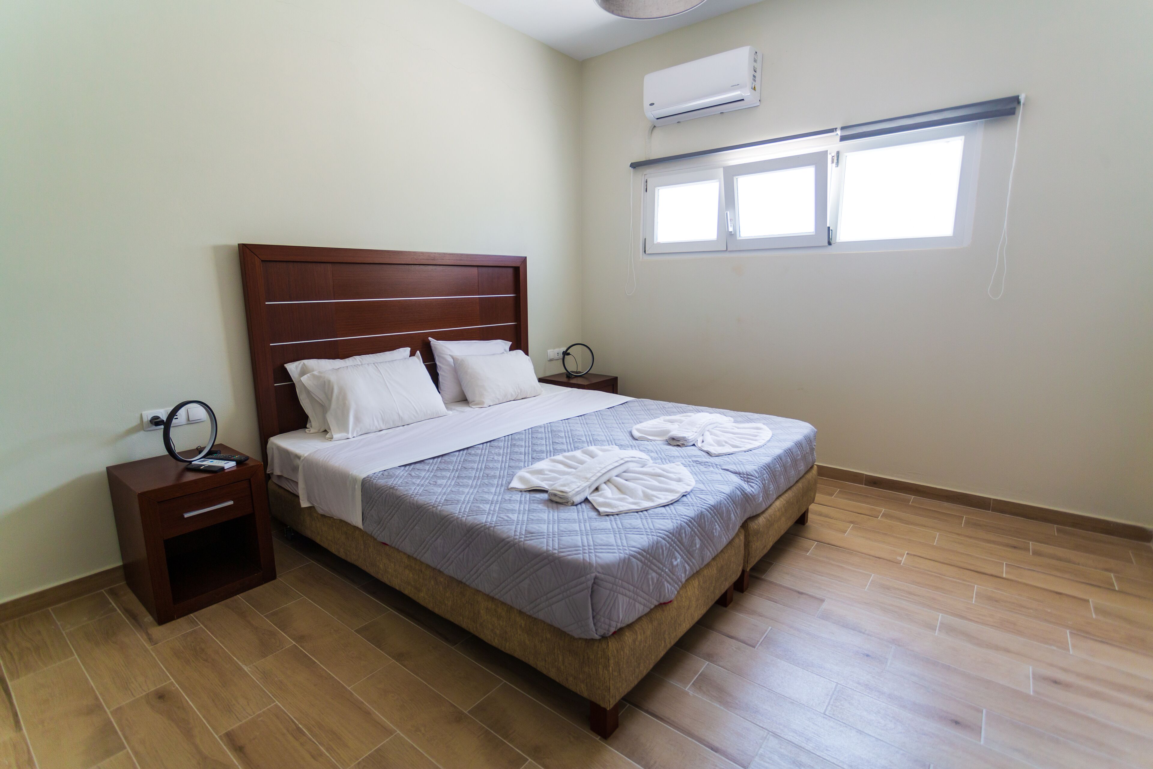 A neatly made bed with white linens and towels, a small bedside table with a ring light, and a window with blinds.